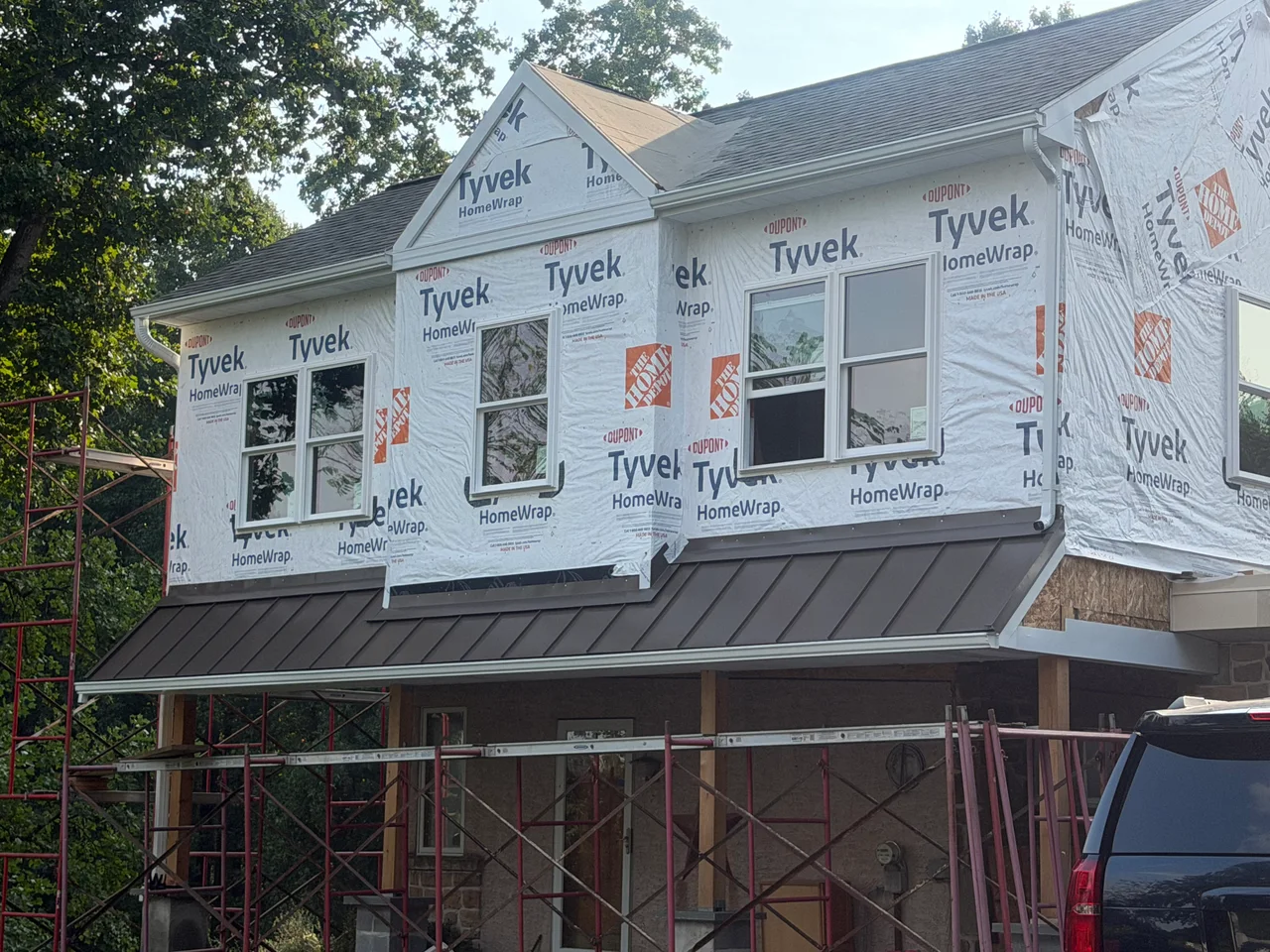 Two-story house exterior with white printed house wrap around installed windows, gray roof shingles, and a dark standing-seam metal porch roof. Exposed wood posts and she